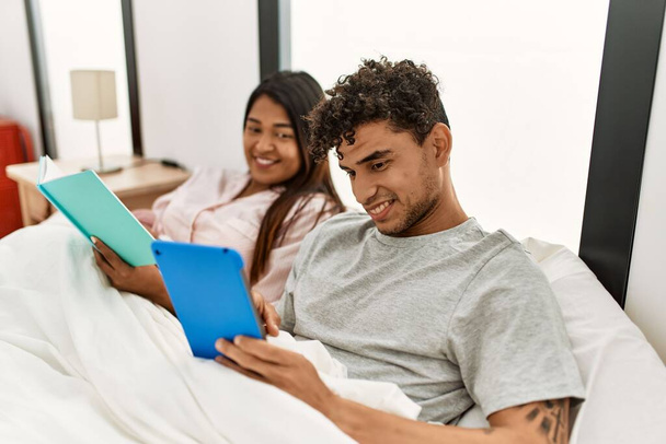 Yatak odasındaki yatakta oturan dokunmatik kâğıt kullanarak kitap okuyan genç Latin çift.. - Fotoğraf, Görsel