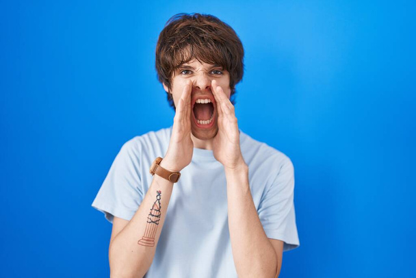 Ισπανόφωνος νεαρός άνδρας στέκεται πάνω από το μπλε φόντο φωνάζοντας θυμωμένος δυνατά με τα χέρια πάνω από το στόμα  - Φωτογραφία, εικόνα