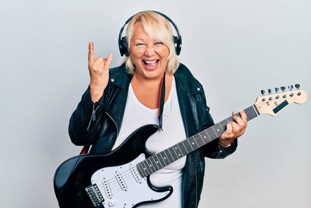Middle age blonde woman playing electric guitar doing horns sign with fingers smiling and laughing hard out loud because funny crazy joke.  - Fotografie, Obrázek