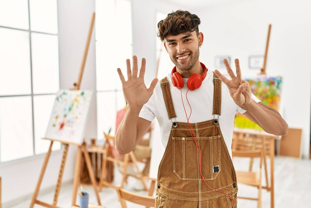 Jeune homme hispanique au studio d'art montrant et pointant vers le haut avec les doigts numéro huit tout en souriant confiant et heureux.  - Photo, image