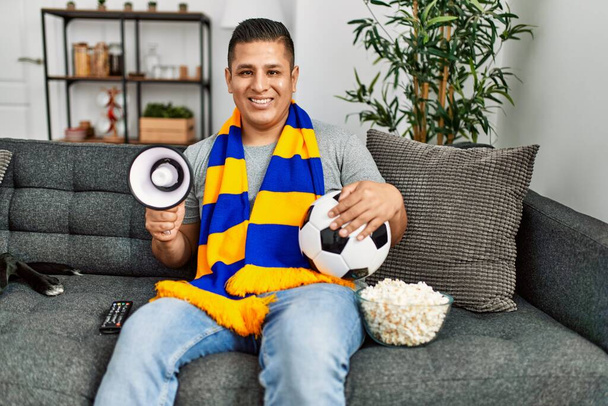 Joven hooligan hispano apoyando al equipo de fútbol sentado en el sofá en casa. - Foto, Imagen