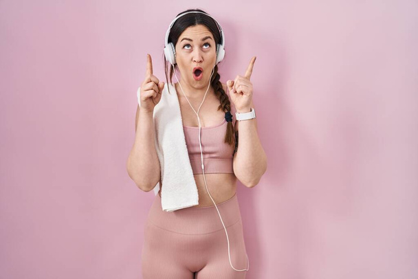 Young brunette woman wearing sportswear and headphones amazed and surprised looking up and pointing with fingers and raised arms.  - Фото, зображення