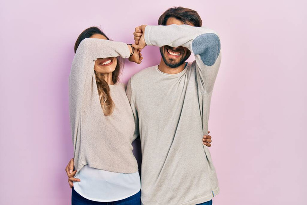 Joven pareja hispana vistiendo ropa casual sonriendo alegre jugando a echar un vistazo a un boo con las manos mostrando la cara. sorprendido y salido  - Foto, imagen