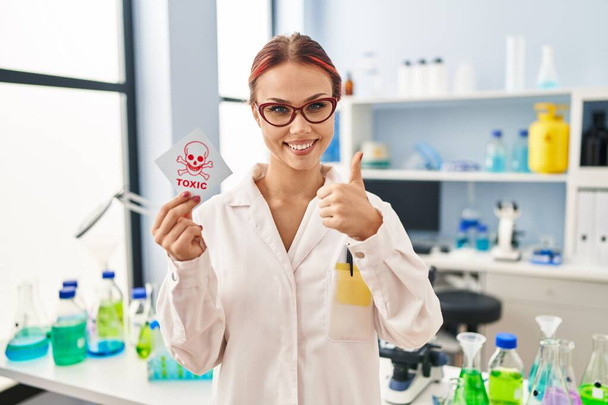 Молодая белая женщина, работающая в научной лаборатории с токсичной этикеткой, улыбаясь счастливой и позитивной, с большим пальцем вверх, отлично справляясь и одобряя знак  - Фото, изображение