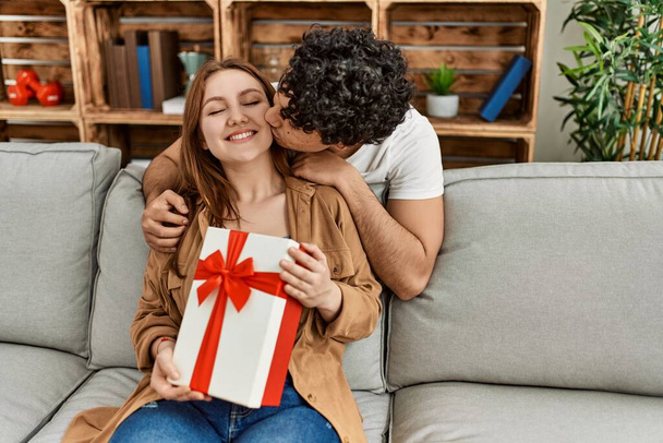Νεαρό ζευγάρι χαμογελά χαρούμενος κάθεται στον καναπέ έκπληξη με δώρο επετείου στο σπίτι. - Φωτογραφία, εικόνα