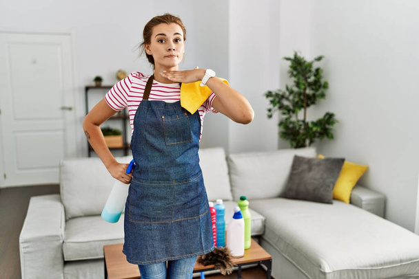 Jeune femme brune portant un tablier tenant des produits de nettoyage à la maison tranchant la gorge avec la main comme couteau, menace d'agression avec une violence furieuse  - Photo, image