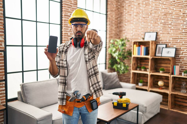 Junger hispanischer Mann mit Bart bei Renovierungsarbeiten zu Hause, Smartphone mit Zeigefinger in die Kamera und auf Sie gerichtet, selbstbewusste Geste, die ernst wirkt  - Foto, Bild