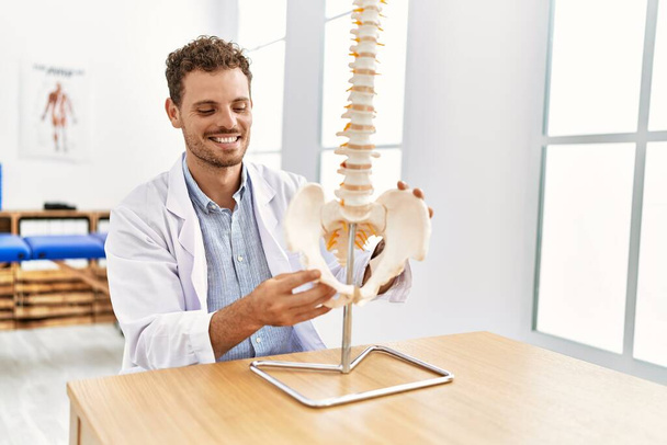 Joven hispano vistiendo uniforme fisioterapeuta tocando modelo anatómico de columna vertebral en clínica - Foto, imagen