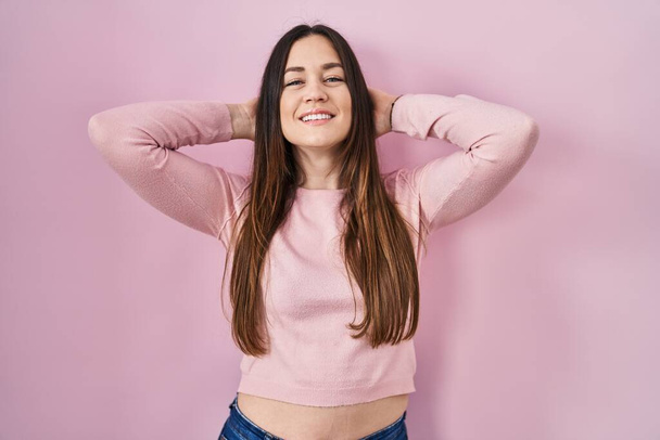 Jeune femme brune debout sur fond rose relaxant et étirant, bras et mains derrière la tête et le cou souriant heureux  - Photo, image