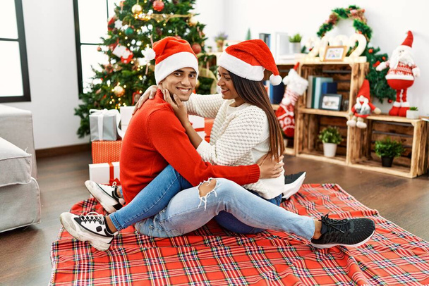 Young latin couple smiling happy and hugging sitting by christmas tree at home. - Photo, Image