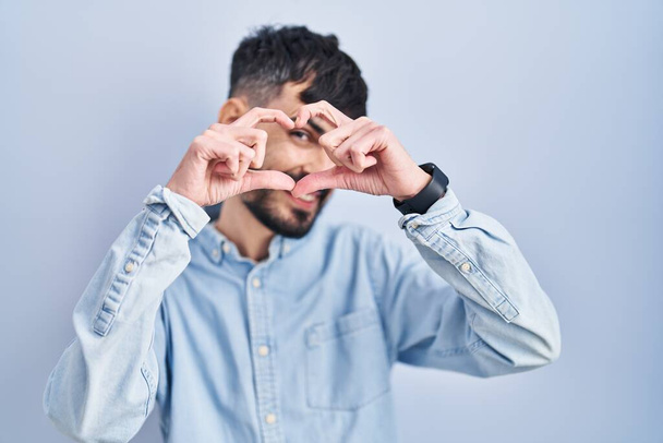 Молодой латиноамериканец с бородой стоит на синем фоне и делает форму сердца с рукой и пальцами, улыбаясь, глядя сквозь знак  - Фото, изображение
