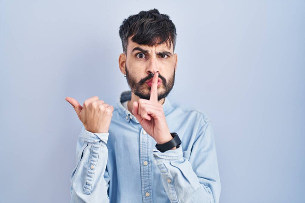Νεαρός Ισπανός με γενειάδα που στέκεται πάνω από μπλε φόντο και ζητάει ησυχία με το δάχτυλο στα χείλη να δείχνει με το χέρι στο πλάι. σιωπή και μυστική έννοια.  - Φωτογραφία, εικόνα