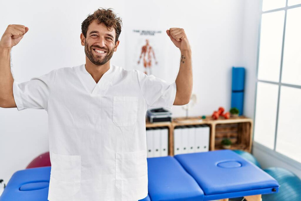 Νεαρός όμορφος φυσιοθεραπευτής που εργάζεται στην κλινική αποκατάστασης πόνου και δείχνει τους μύες των χεριών να χαμογελούν υπερήφανοι. έννοια της καταλληλότητας.  - Φωτογραφία, εικόνα