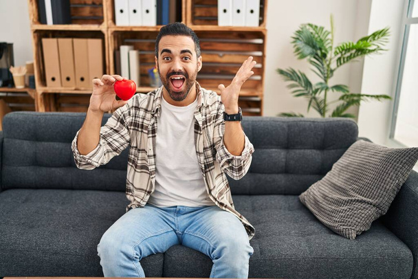 Young hispanic man with beard working on love therapy at consultation office celebrating victory with happy smile and winner expression with raised hands  - Foto, afbeelding