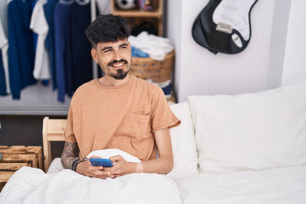 Young hispanic man using smartphone sitting on bed at bedroom - Foto, Imagen