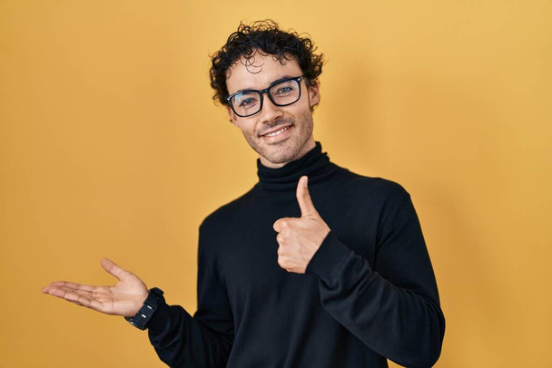 Hispanic man standing over yellow background showing palm hand and doing ok gesture with thumbs up, smiling happy and cheerful  - Foto, afbeelding