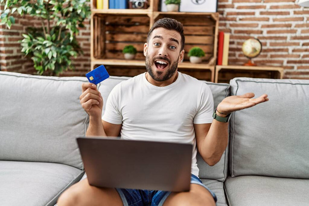 Jóképű spanyol férfi csinál online vásárlás holding hitelkártya ünneplő teljesítmény boldog mosollyal és a győztes kifejezés emelt kézzel  - Fotó, kép