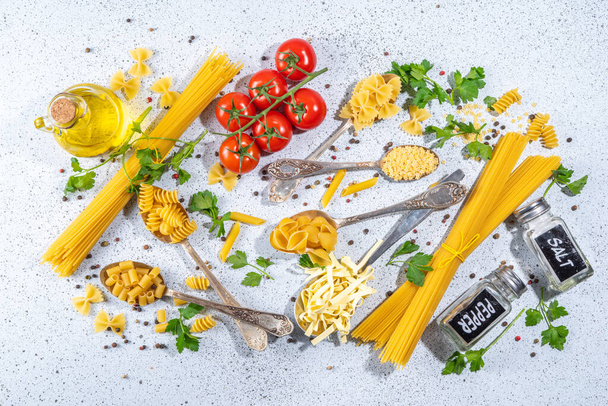 様々な乾燥スパゲティ、麺、白いテーブルの背景にスプーンで設定されたパスタ。ハーブ、オリーブオイル、トマト、スパイスをトップビューのコピースペースを調理するための異なる形状の生のイタリアのパスタ - 写真・画像
