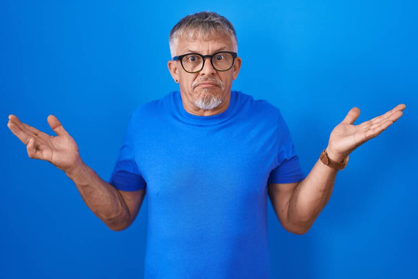 Hispanic man with grey hair standing over blue background clueless and confused expression with arms and hands raised. doubt concept.  - Photo, Image