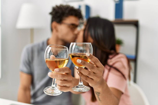 Junges lateinisches Paar küsst und stößt mit Wein an, der zu Hause auf dem Tisch liegt. - Foto, Bild