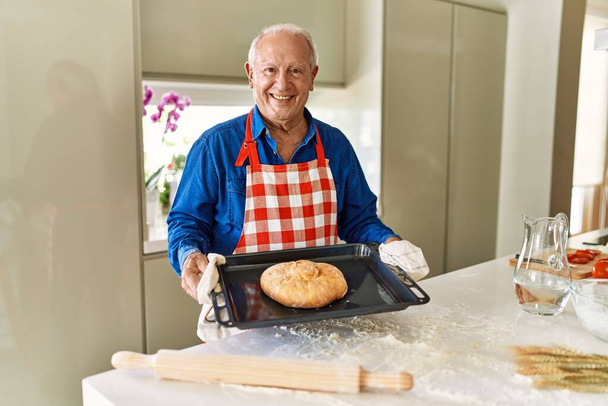 Uomo anziano che tiene il vassoio del forno con pane fatto in casa in cucina - Foto, immagini