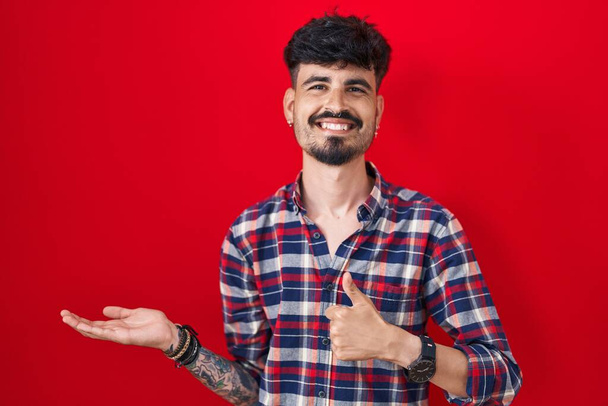 Young hispanic man with beard standing over red background showing palm hand and doing ok gesture with thumbs up, smiling happy and cheerful  - Photo, Image