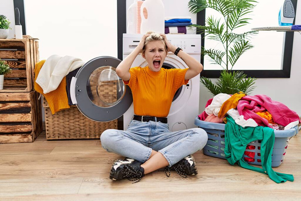 Молодая блондинка, занимающаяся стиркой, сидя у стиральной машины сумасшедшая и напуганная руками на голове, напуганная и удивленная шоком с открытым ртом  - Фото, изображение