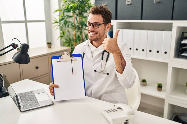Junger hispanischer Mann im Doktor-Stethoskop mit Klemmbrett, der glücklich und positiv lächelt, Daumen hoch und Zeichen der Anerkennung  - Foto, Bild