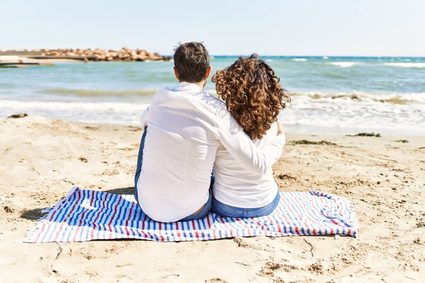 Middle age hispanic couple on back view sitting on the towel hugging at the beach. - Photo, Image