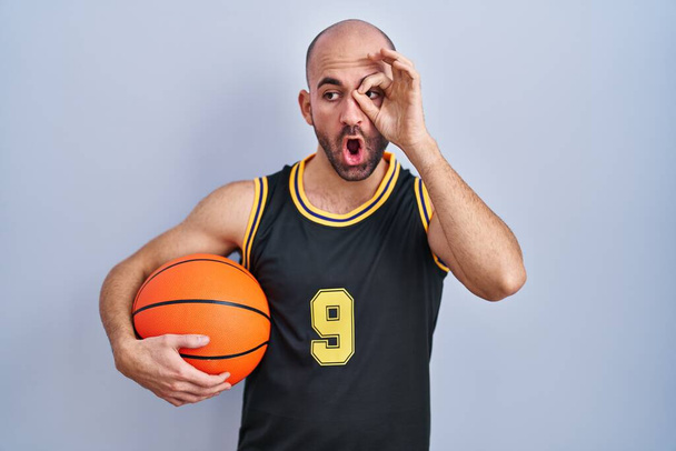Joven calvo con barba vistiendo uniforme de baloncesto sosteniendo la pelota haciendo buen gesto sorprendido con la cara sorprendida, los ojos mirando a través de los dedos. expresión incrédula.  - Foto, imagen