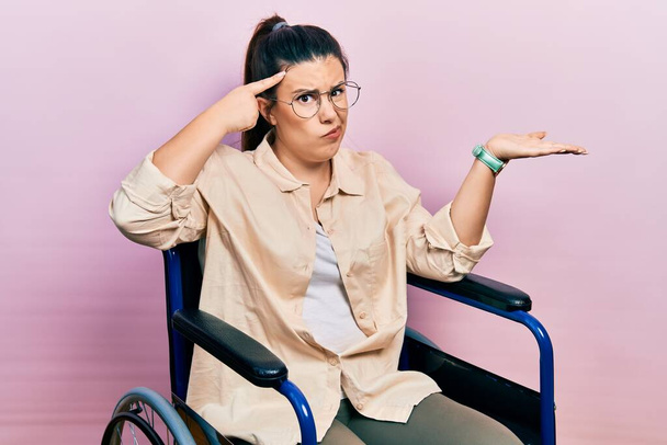 Young hispanic woman sitting on wheelchair confused and annoyed with open palm showing copy space and pointing finger to forehead. think about it.  - Foto, afbeelding