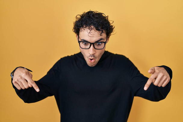 Hispanic man standing over yellow background pointing down with fingers showing advertisement, surprised face and open mouth  - Foto, afbeelding