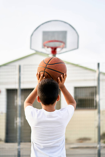 A cute young boy plays basketball on the street playground in summer. Teenager in a white t-shirt with orange basketball ball outside. Hobby, active lifestyle, sports activity for kids. - Fotoğraf, Görsel