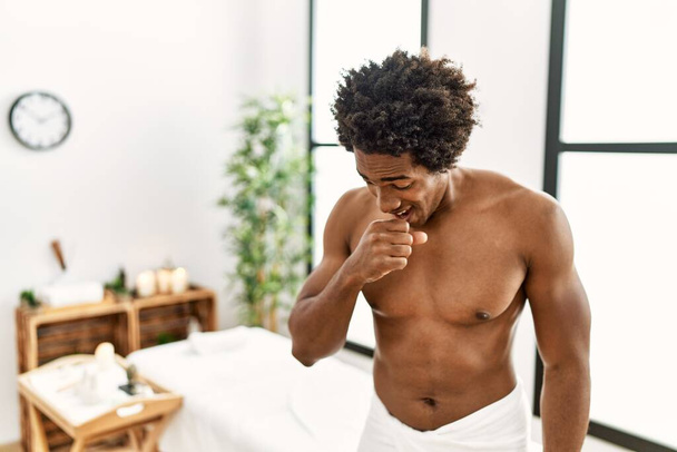Jonge Afro-Amerikaanse man zonder shirt die een handdoek draagt terwijl hij in het schoonheidscentrum staat en zich onwel voelt en hoest als symptoom voor verkoudheid of bronchitis. Gezondheidszorgconcept.  - Foto, afbeelding