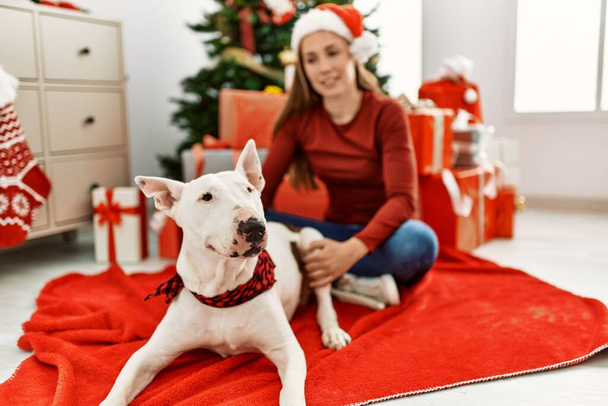 Young caucasian woman smiling confident sitting with dog by christmas tree at home - Photo, Image