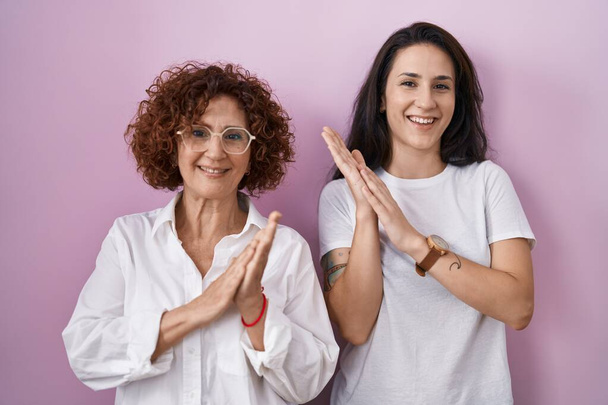 Hispanic mother and daughter wearing casual white t shirt over pink background clapping and applauding happy and joyful, smiling proud hands together  - Photo, Image