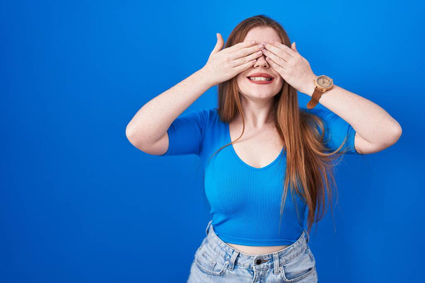 Redhead woman standing over blue background covering eyes with hands smiling cheerful and funny. blind concept.  - Photo, Image