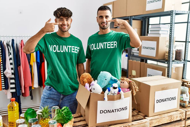 Νεαρό γκέι ζευγάρι που φοράει εθελοντικό μπλουζάκι σε δωρεές στέκεται δείχνοντας με το δάχτυλο στο πρόσωπο και τη μύτη, χαμογελώντας χαρούμενα. έννοια ομορφιά  - Φωτογραφία, εικόνα