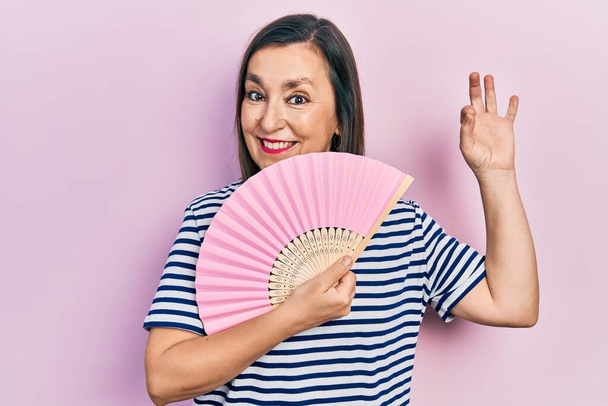 Middle age hispanic woman waving hand fan cooling air in summer doing ok sign with fingers, smiling friendly gesturing excellent symbol  - Foto, Bild