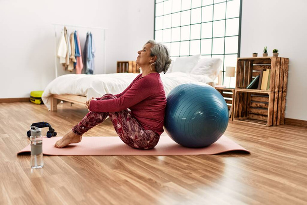 Середнього віку сіро-волохата жінка тренується з підходящим м'ячем, сидячи на підлозі в спальні
. - Фото, зображення