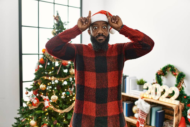 Αφροαμερικάνος που φοράει το καπέλο του Άη Βασίλη στέκεται δίπλα στο χριστουγεννιάτικο δέντρο κάνοντας αστεία χειρονομία με δάχτυλο πάνω από το κεφάλι σαν κέρατα ταύρου  - Φωτογραφία, εικόνα