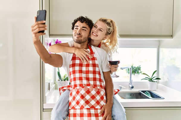 Mladý pár se šťastně objímal a seděl na pultě. Pití červeného vína a používání smartphonu v kuchyni. - Fotografie, Obrázek