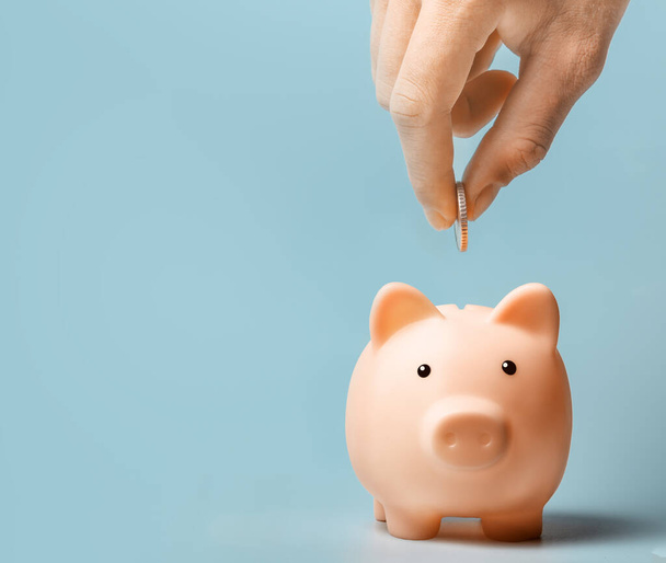 Hand putting coin to pink piggy bank on a blue background. Copy space - Photo, image