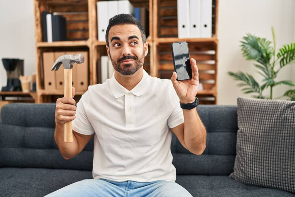 Young hispanic man with beard holding hammer and broken smartphone showing cracked screen smiling looking to the side and staring away thinking.  - Φωτογραφία, εικόνα