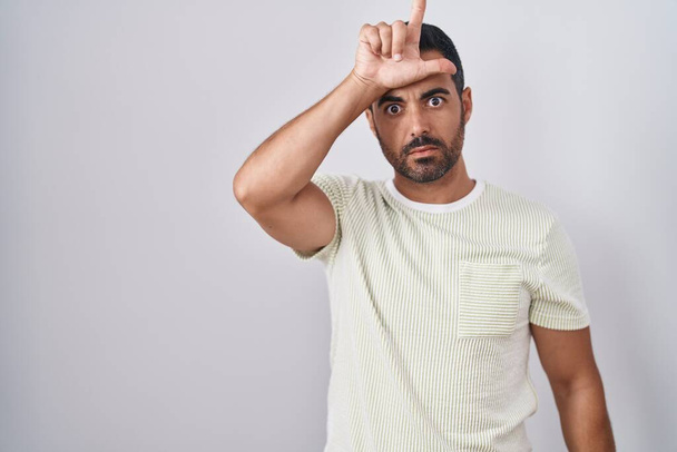 Spaanse man met baard die over een geïsoleerde achtergrond staat en grapjes maakt over mensen met vingers op het voorhoofd die verliezers bespotten en beledigen.  - Foto, afbeelding
