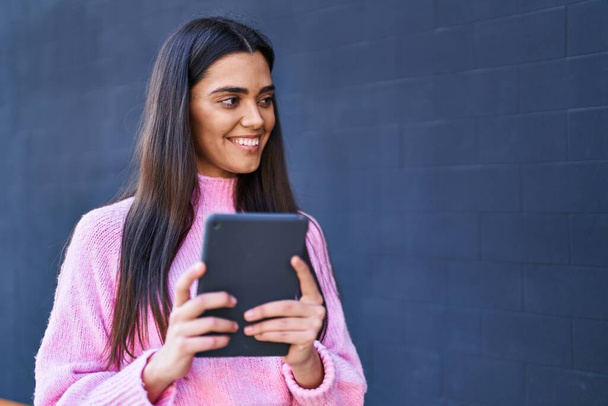 Νεαρή ισπανόφωνη γυναίκα χαμογελά με αυτοπεποίθηση χρησιμοποιώντας touchpad στο δρόμο - Φωτογραφία, εικόνα