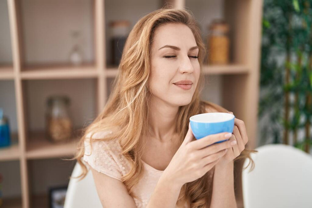 Young blonde woman drinking coffee sitting on table at home - Foto, Imagem