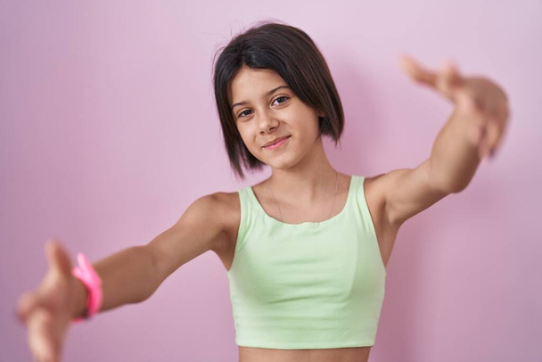 Young girl standing over pink background looking at the camera smiling with open arms for hug. cheerful expression embracing happiness.  - Foto, Imagen