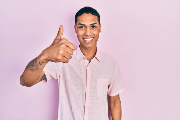 Młody Afroamerykanin ubrany w luźne ubrania uśmiechnięty szczęśliwy i pozytywny, kciuk do góry robi doskonałe i znak aprobaty  - Zdjęcie, obraz