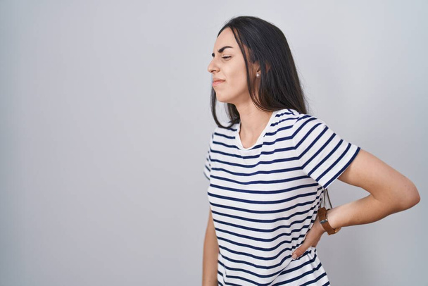Young brunette woman wearing striped t shirt suffering of backache, touching back with hand, muscular pain  - Фото, изображение